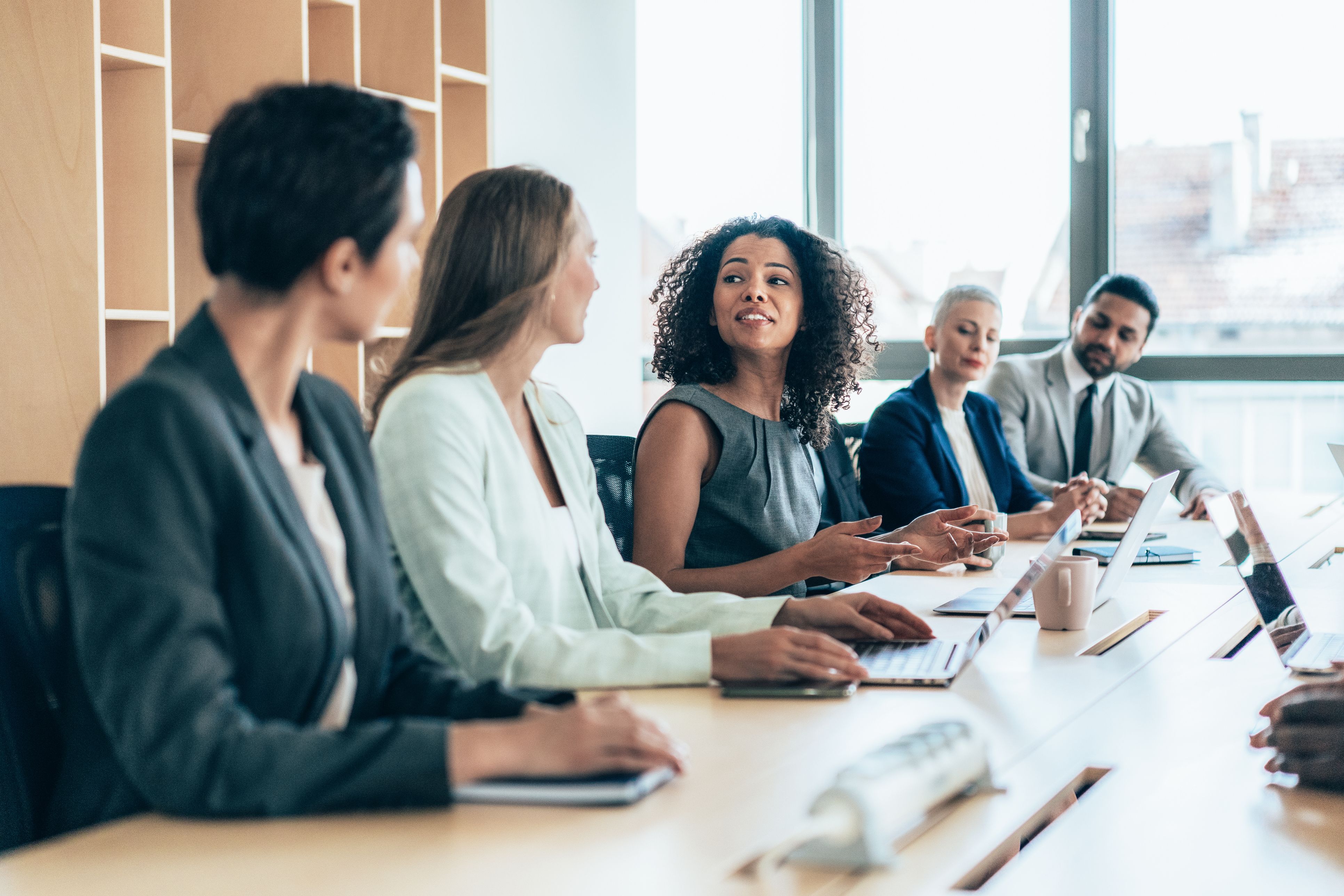 Image of a business meeting with a view down one side of the table. A woman with curly hair is talking and the other four people are looking at her