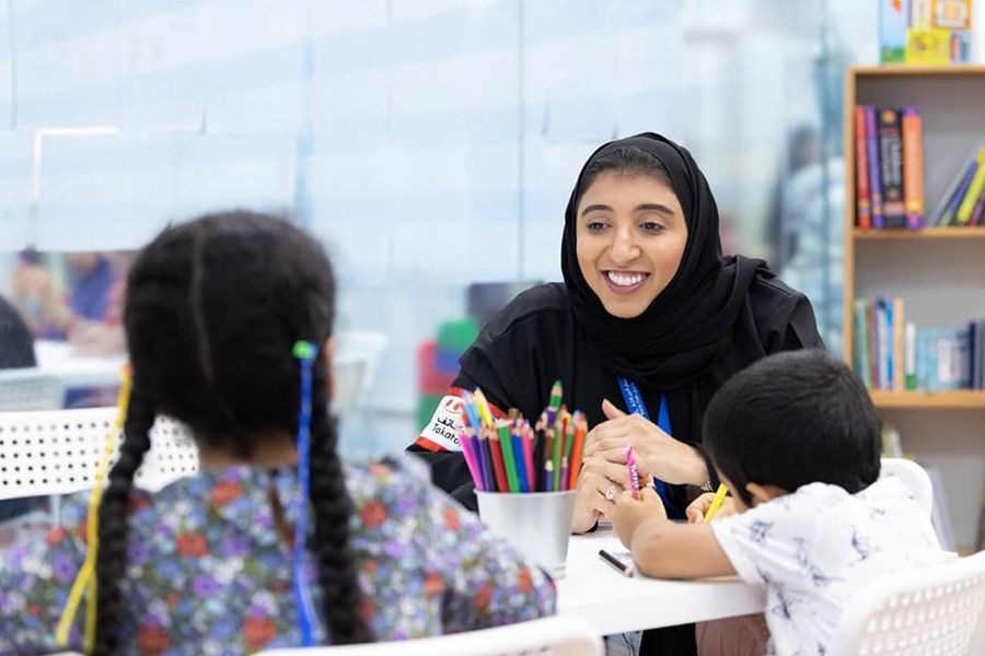 Image showing Eiman Aldarmaki sitting at an arts table smiling and chatting to two small children  (a girl and boy) who are using colouring pencils and have their backs to the camera