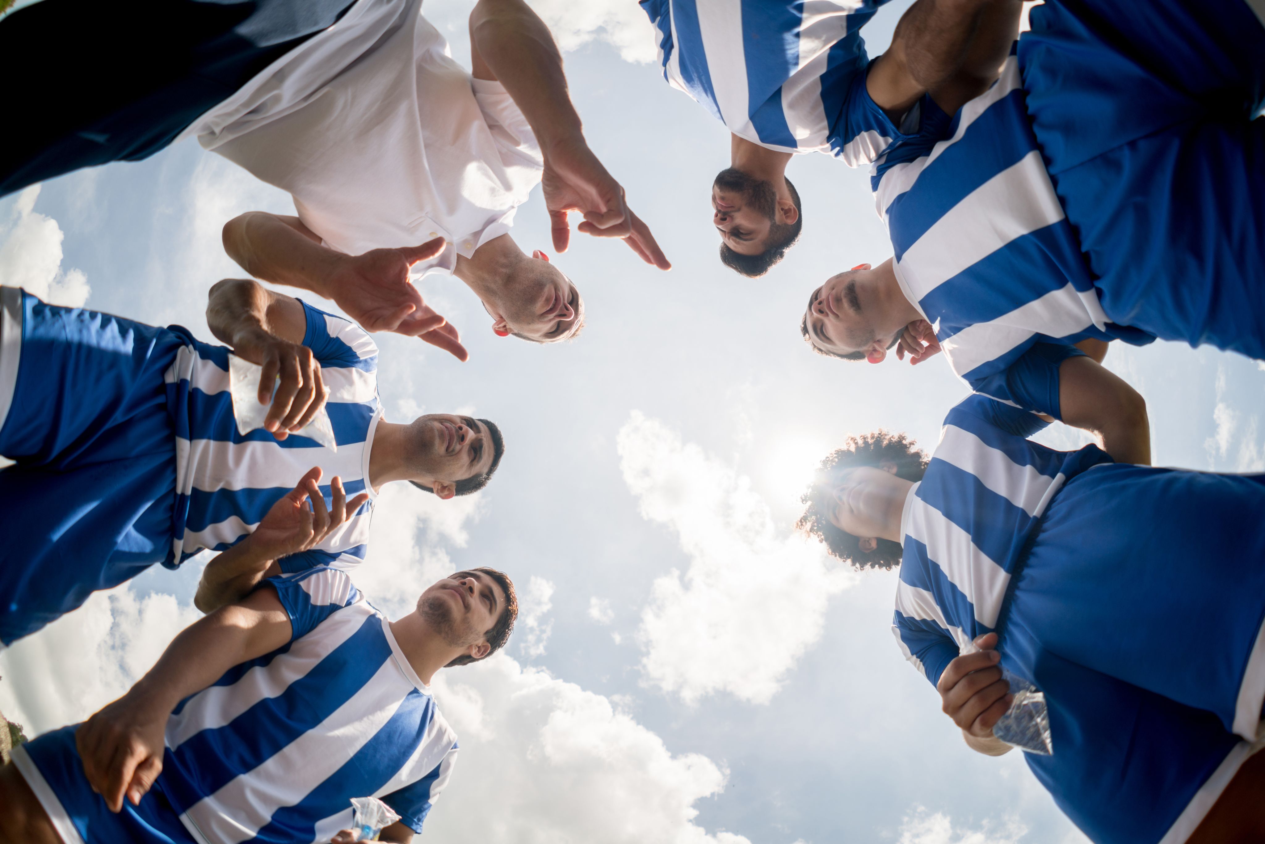 Image of a men's football team dressed in blue and white kit gathered round in a circle with the camera in the middle looking up at them