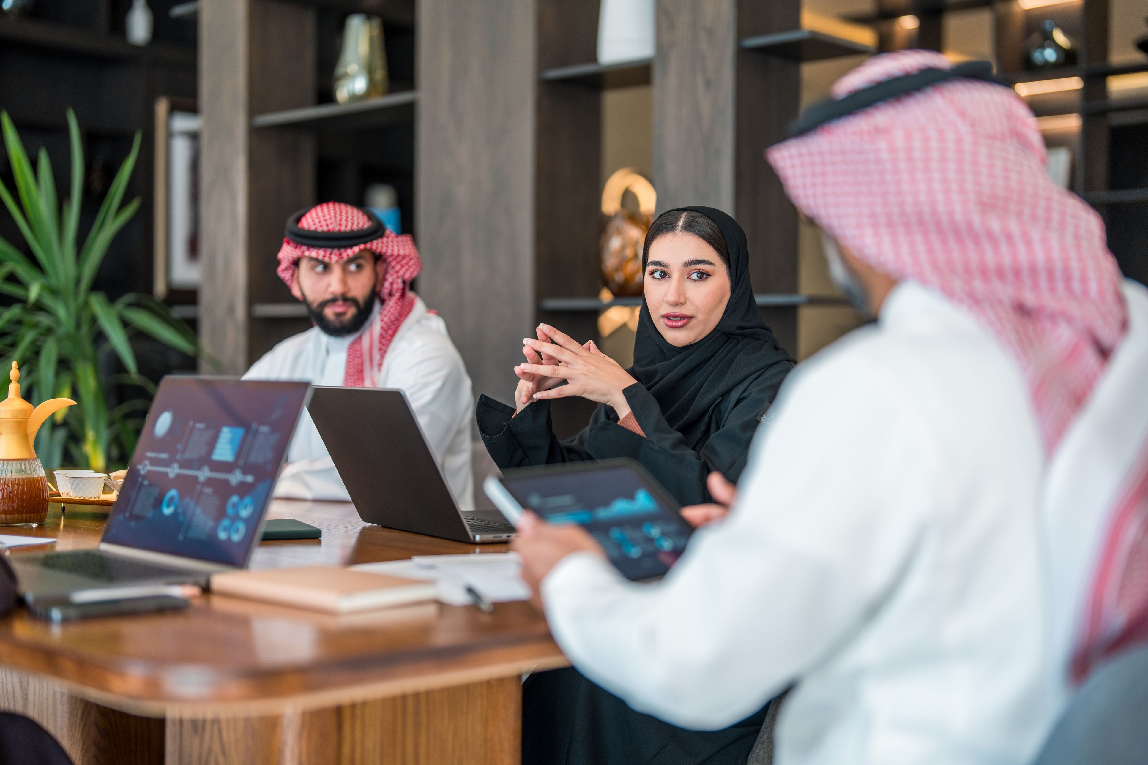 Business executives in Saudi Arabia at a meeting. The man wears traditional attire, while the woman is in a black abaya. Laptops and documents are present in a modern office setting