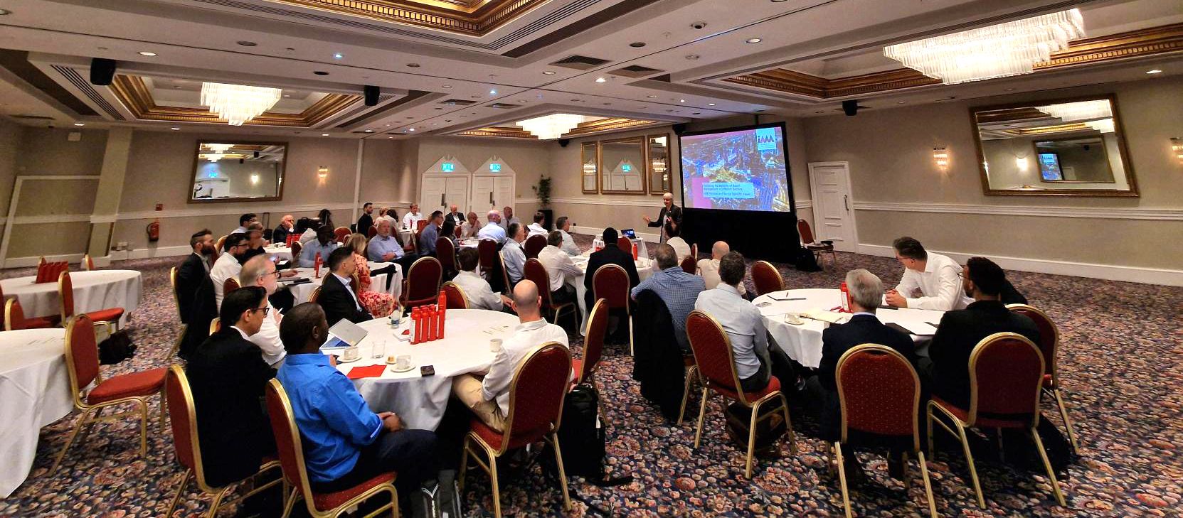 Conference room filled with people sitting at tables and watching an address on screen